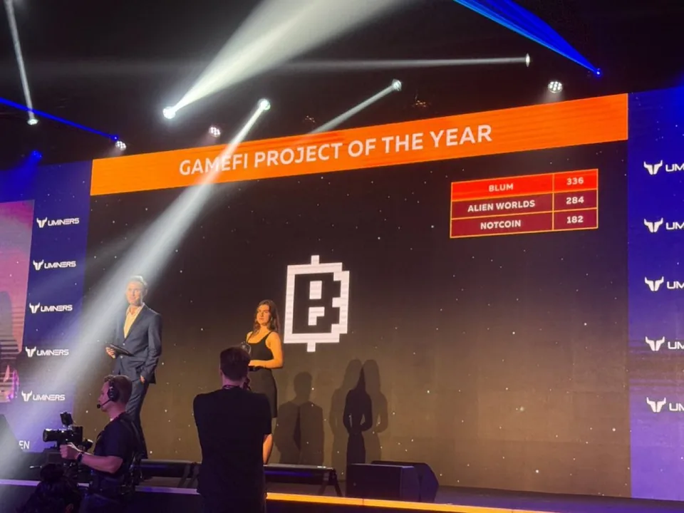 An awards stage: a screen reads GameFi project of the year, with the vote Blum 336, Alien Worlds 284, Notcoin 182, and the Blum pixel B large in the middle; two presenters stand below it.